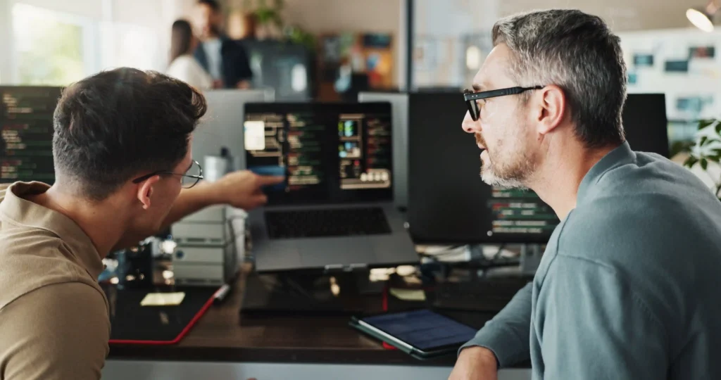 Two coworkers review code on a laptop at a desk with multiple monitors in a modern office.